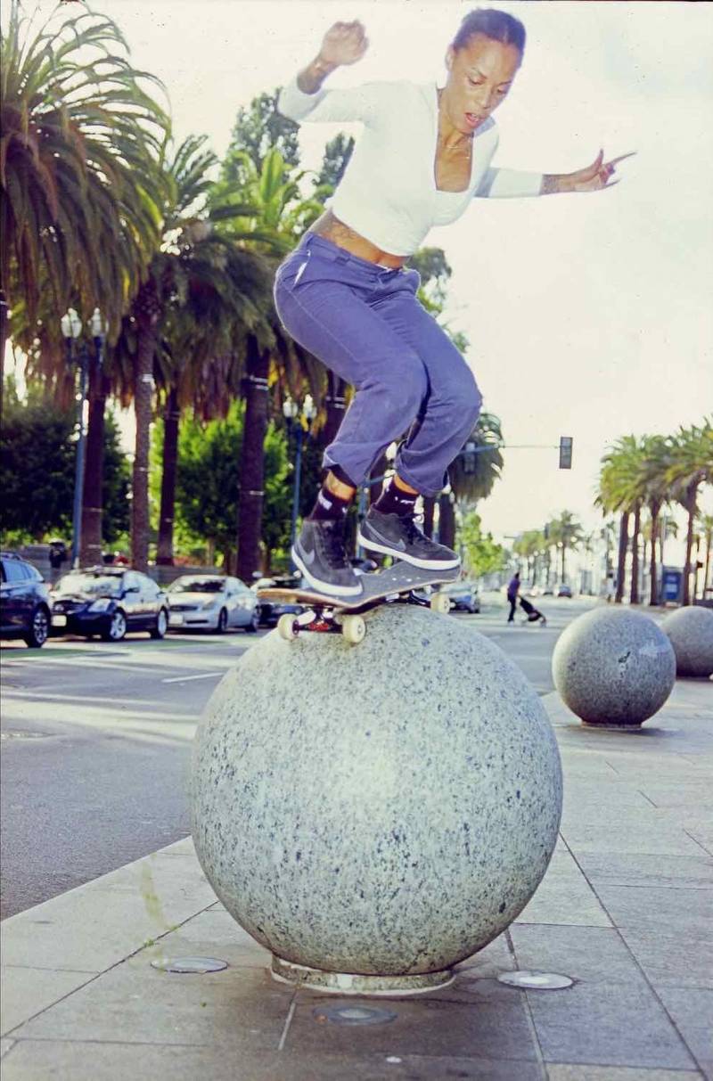 Marsha Howard balances on her skateboard atop a giant cement ball. Palm trees are in the background.