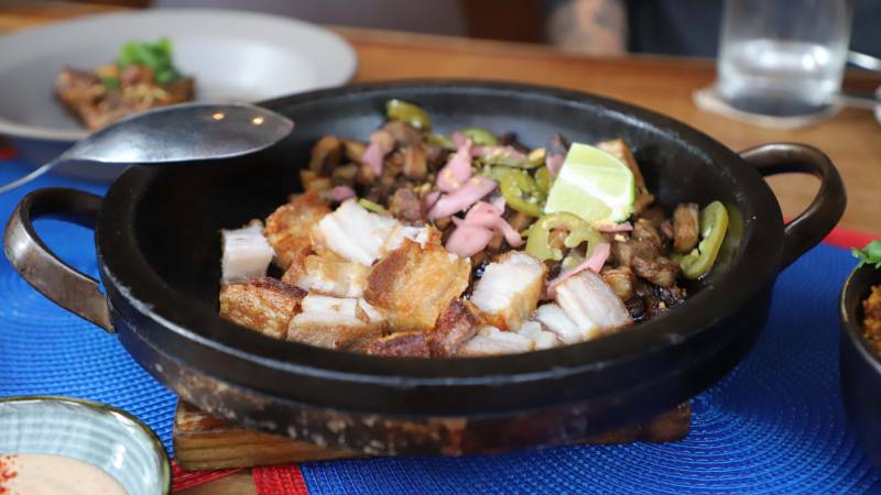 Crispy pork belly (lechon kawali) in a black skillet, topped with lime and pickled onions and jalapeños.