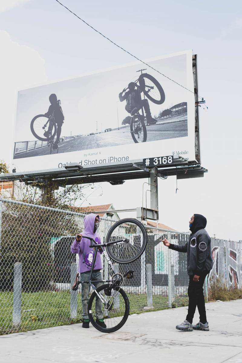 Omar "Meez" Jones and Pershanté Hill standing on 7th Street in West Oakland, talking to each other in front of a billboard that features Jones' image.