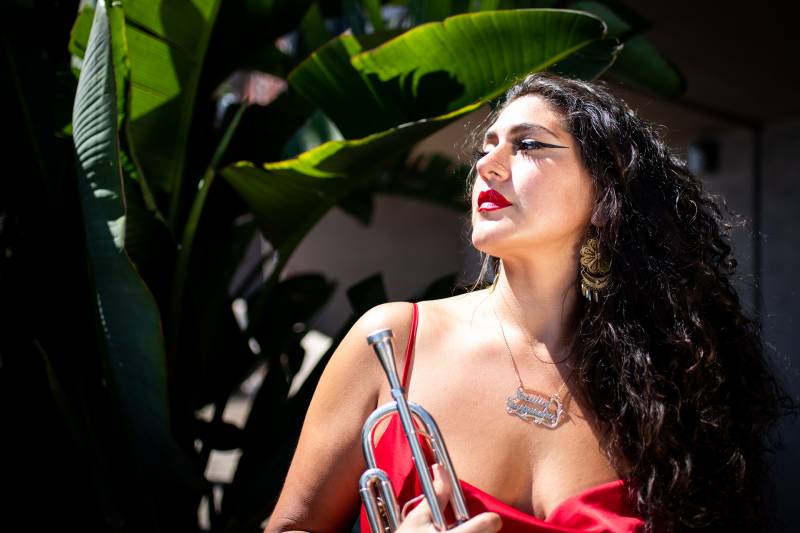 A Mexican American woman in a red dress and black eyeline holds a trumpet among tropical plants.