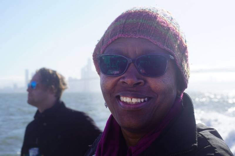 Dorothy Lazard wearing a knit cap and sunglasses, on a boat, with the SF skyline in the background