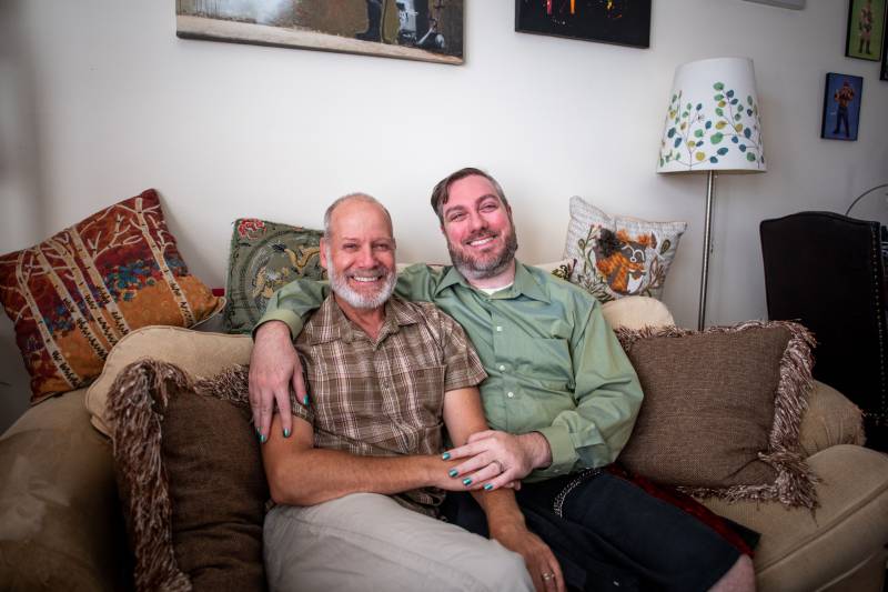 Ron (left) and Erik (right) hold each other, sitting on a couch in their home while posing for a photo.