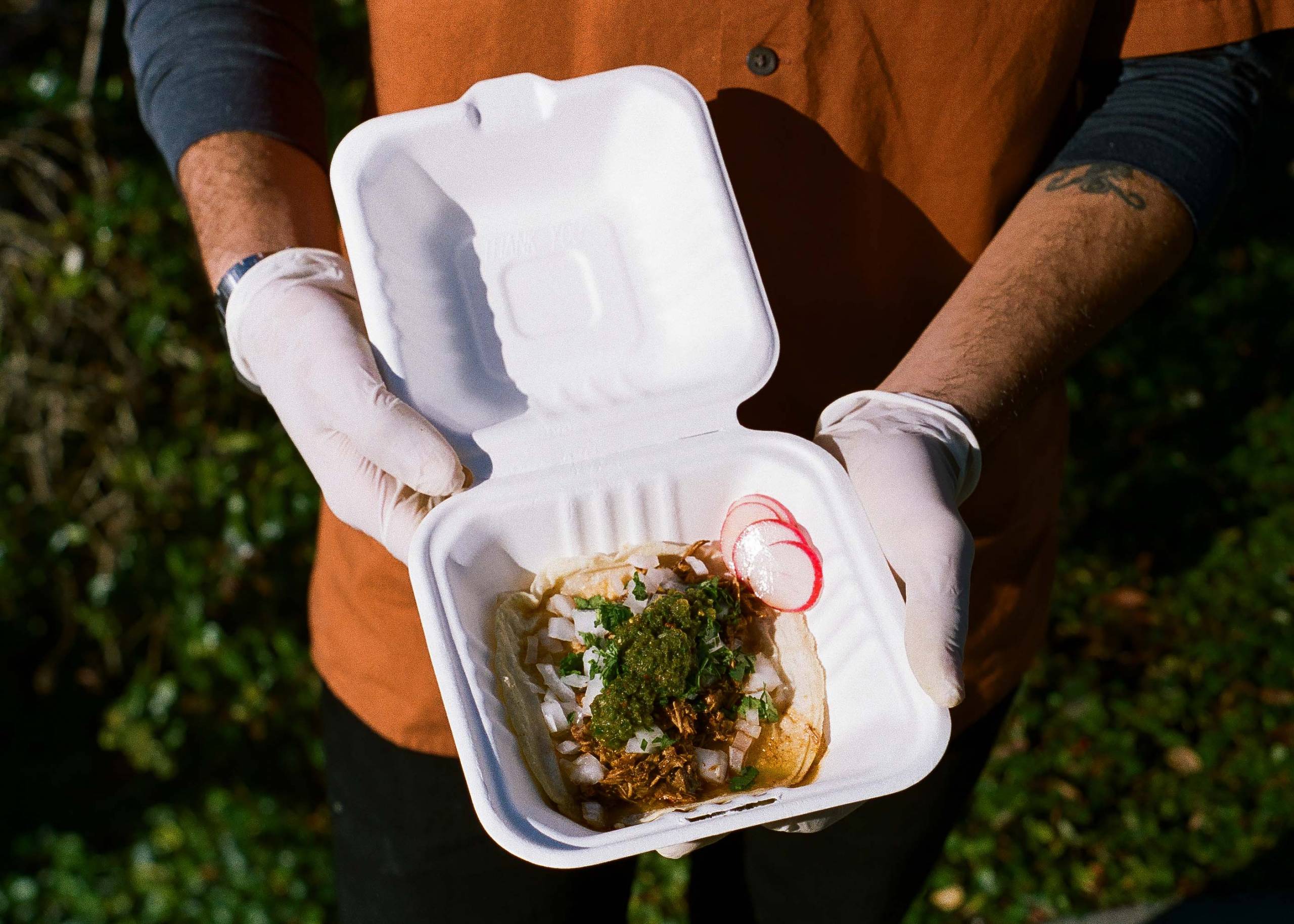 Gloved hands holding a takeout container with a single taco inside.