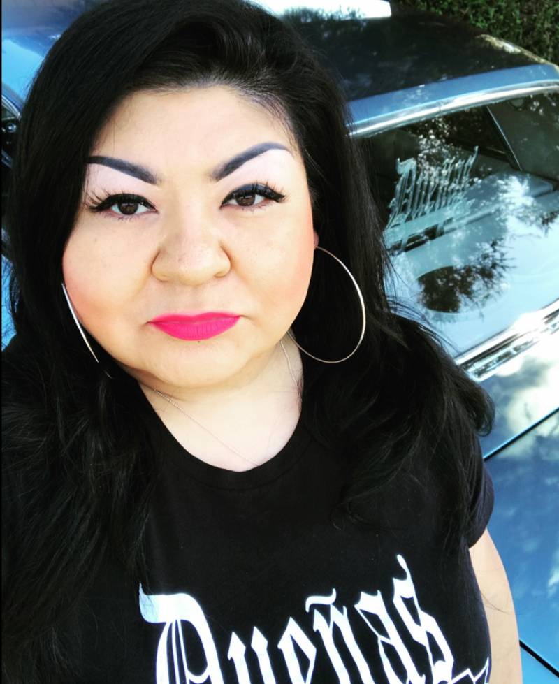 Angel Romero, the founder of the Dueñas Car Club, takes a selfie in front of her car, through the window you can see the Dueñas logo, it's also on her black t-shirt.