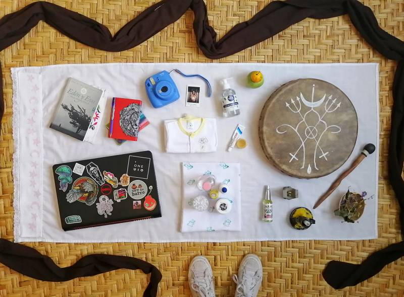 Items, left to right: Book, notebooks, laptop, instant camera with print, baby clothes, personal hygiene supplies for mother and baby, hand sanitizer, local fruits, ointment, shamanic drum, Agua de Florida ritual water, prayer altar, spiritual candle, herbs. Also pictured: a black baby wrap surrounds the items with the photographer's shoes visible.