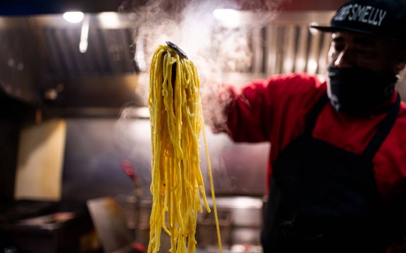 A chef holding a large tangle of steaming garlic noodles with a pair of tongs.