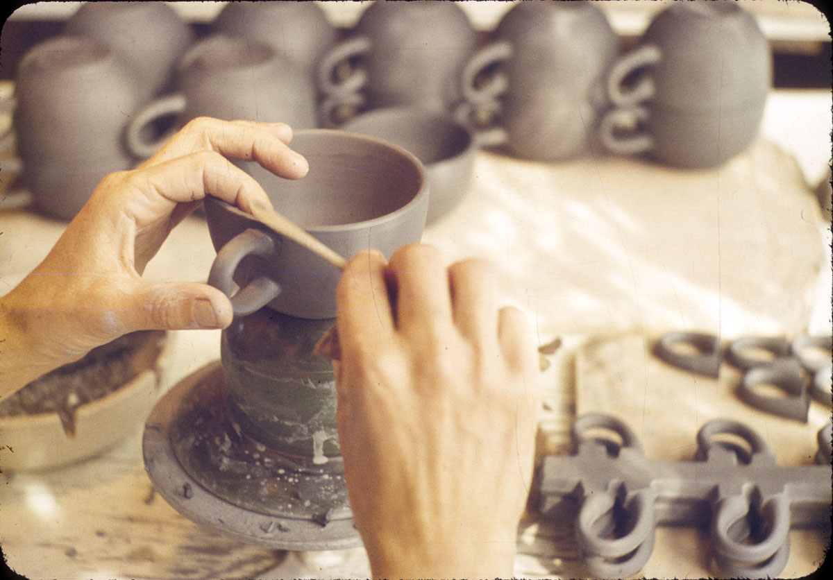 Fair-skinned hands use a tool on a dark clay teacup.