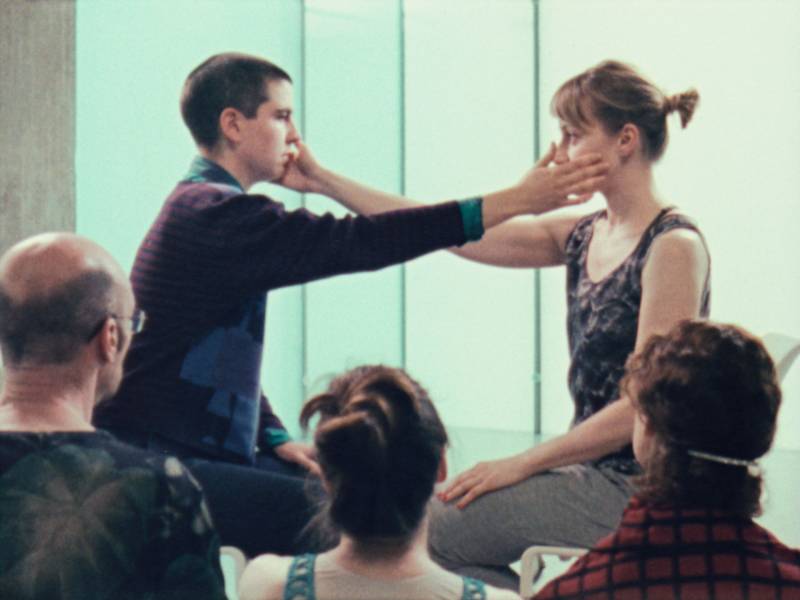 Two white people sit facing each other on their heels in front of a three-person audience. They each reach their right arm out to touch each others left cheek.