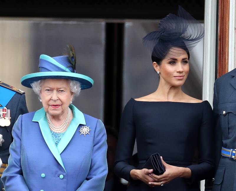 Queen Elizabeth, wearing a lilac and green suit and hat, stands beside Meghan Markle, who's wearing a navy dress and matching fascinator.