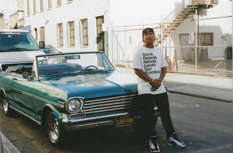 Harvey Lozada stands in front of a classic American made car with a personalized plate that reads "San Fran," while wearing a shirt that reads "Born & Raised & Native & Local & San Franciscan."