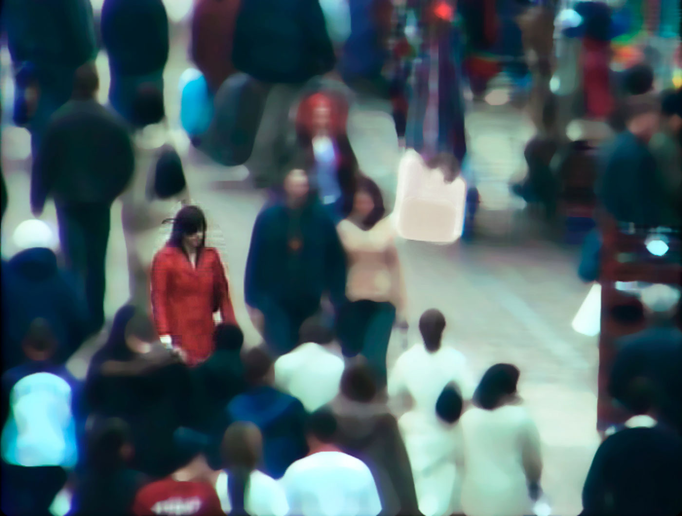 A dark-haired woman in a red coat stands in a crowded street. Only her image is crisp, the other figures have been blurred.