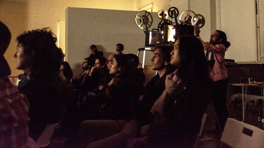 A seated crowd looks left, behind them a projectionists stands next to two film projectors, one actively beaming light.