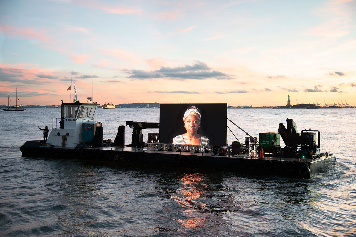 A screen mounted to a floating barge shows a portrait of a woman.