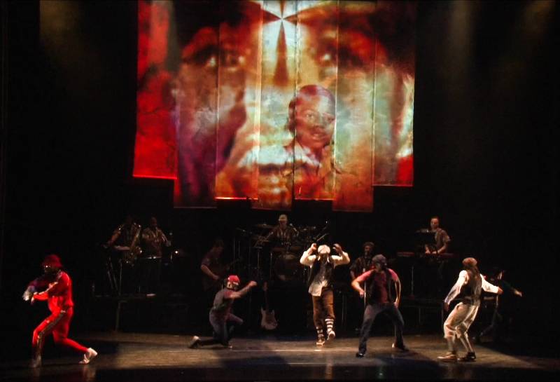 A still from a hip-hop theater production featuring several dancers on a stage of a theater with a big projection screen showing childhood photos of Rennie Harris.