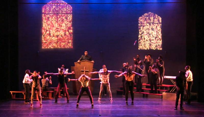 A still from a hip-hop theater performance that shows dancers and singers on a stage in front of an actor playing a priest.