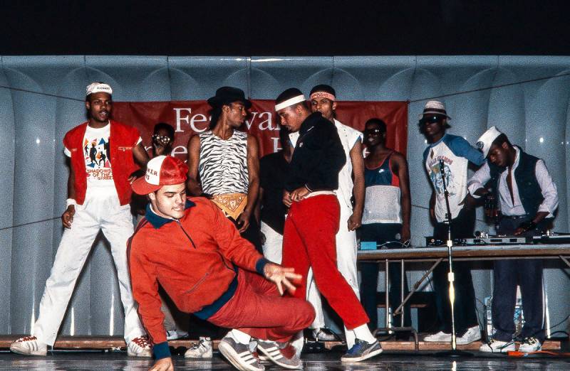 A hip-hop dance and dj crew in the 1980s featuring young male dancers performing on stage while the djs in the back get ready to play a record.