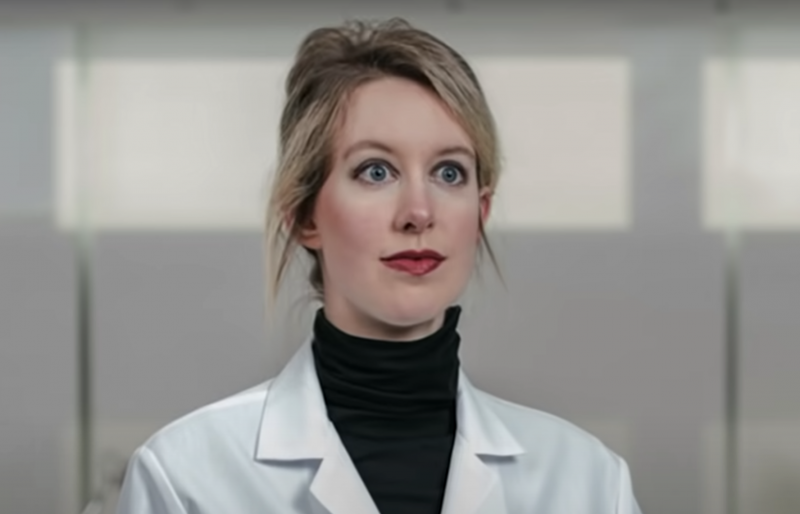 Elizabeth Holmes stares off into the distance with wide blue eyes, hair pulled back in a messy bun, and wearing a lab coat with a black turtleneck underneath.