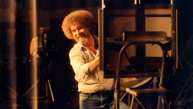 Bob Ross peaks out and smiles from behind a canvas he is painting.