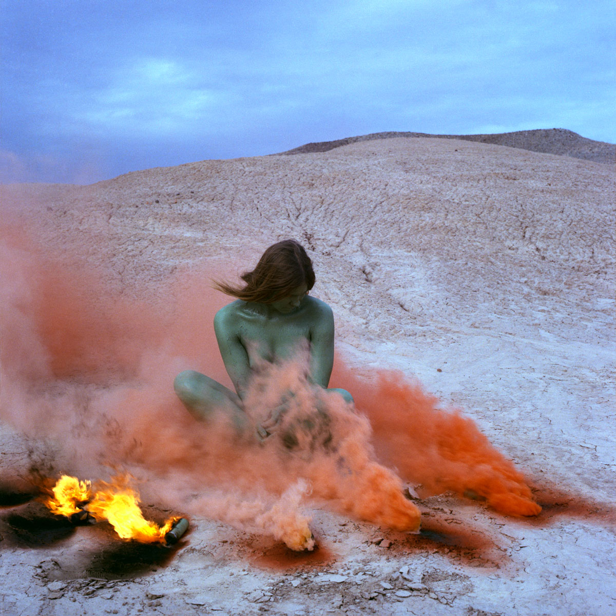 Woman with green skin sits in a cloud of orange smoke.
