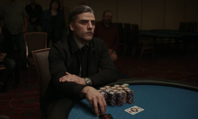 A sharply dressed man sits at a green felt table, many stacks of gambling chips lined up before him.