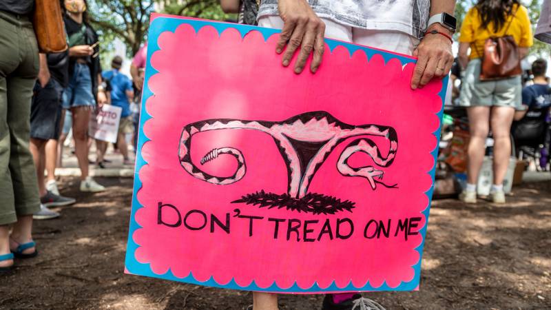 Hands hold a bright pink sign of a uterus that reads "DON'T TREAD ON ME."