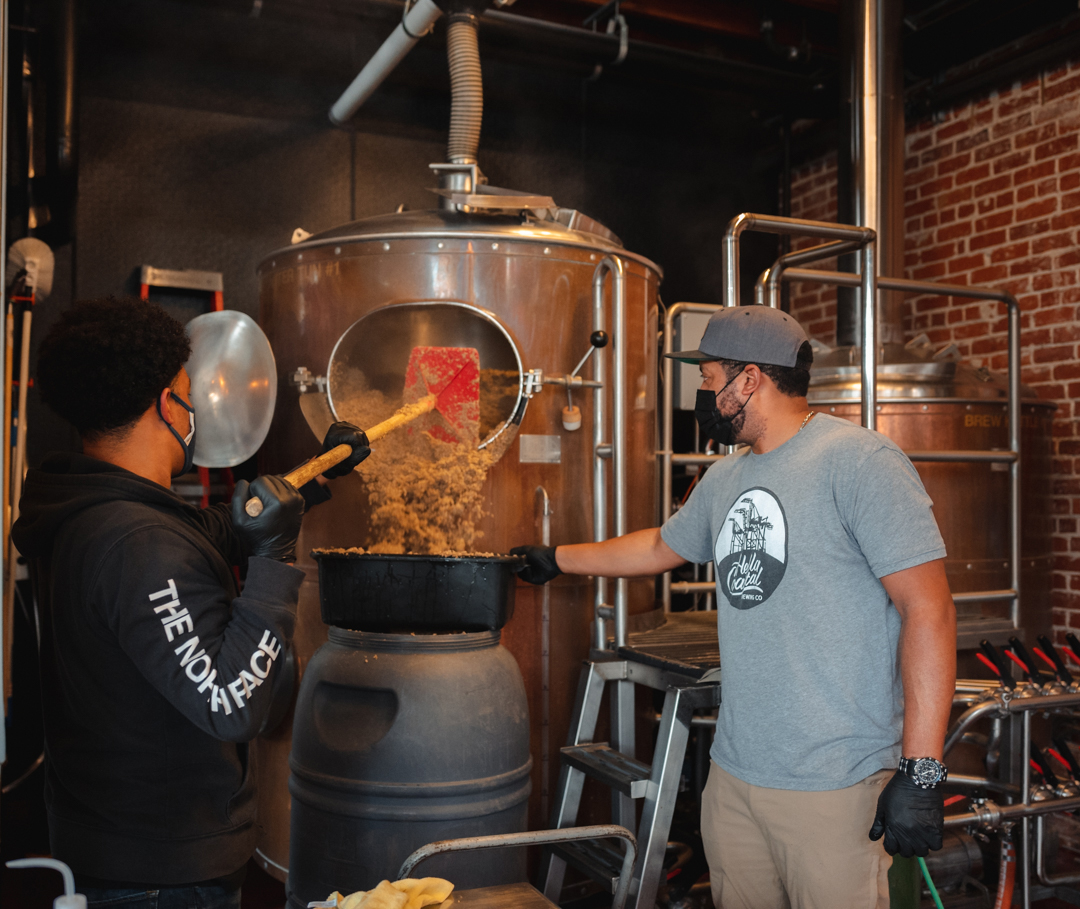 Two men making beer with professional brewing equipment; one wears a visor for protection.