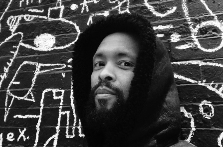 In a black and white photo, Tajai Massey posses in a black hoodie in front of an artistic background that shows what appear to be painted squiggles and designs