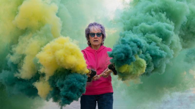 Older woman holds sticks with green and yellow smoke coming out.