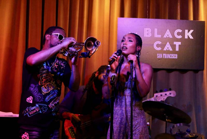 A man in sunglasses plays the trumpet while a woman sings into a microphone.