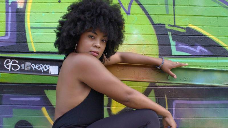 Porshia Derival sports an afro hairstyle and looks straight into the camera while posing in front of a bright green graffiti wall in New York City.