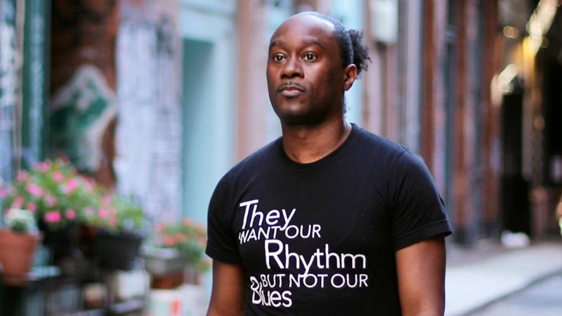 African American man wearing a black t-shirt with white text that reads “They want our rhythm, but not our blues.” 