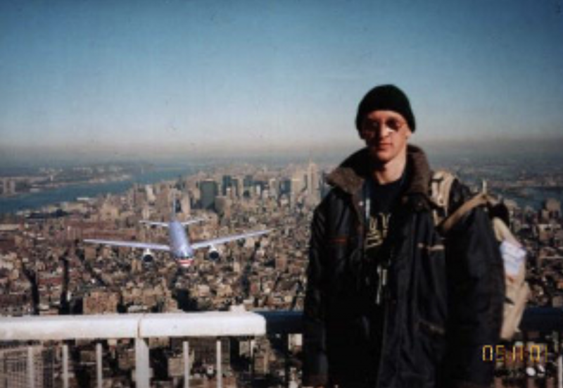 A man stands on the World Trade Center's observation deck with New York City's skyline behind him. In the left of the photo is an approaching commercial airplane. 