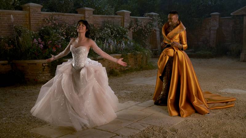 Cinderella, in a white tulle dress stands with arms outstretched as Fab G, wearing a gold formal gown, proudly watches on, smiling.