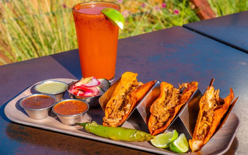 A michelada and a tray of three quesabirria tacos on an outdoor table.