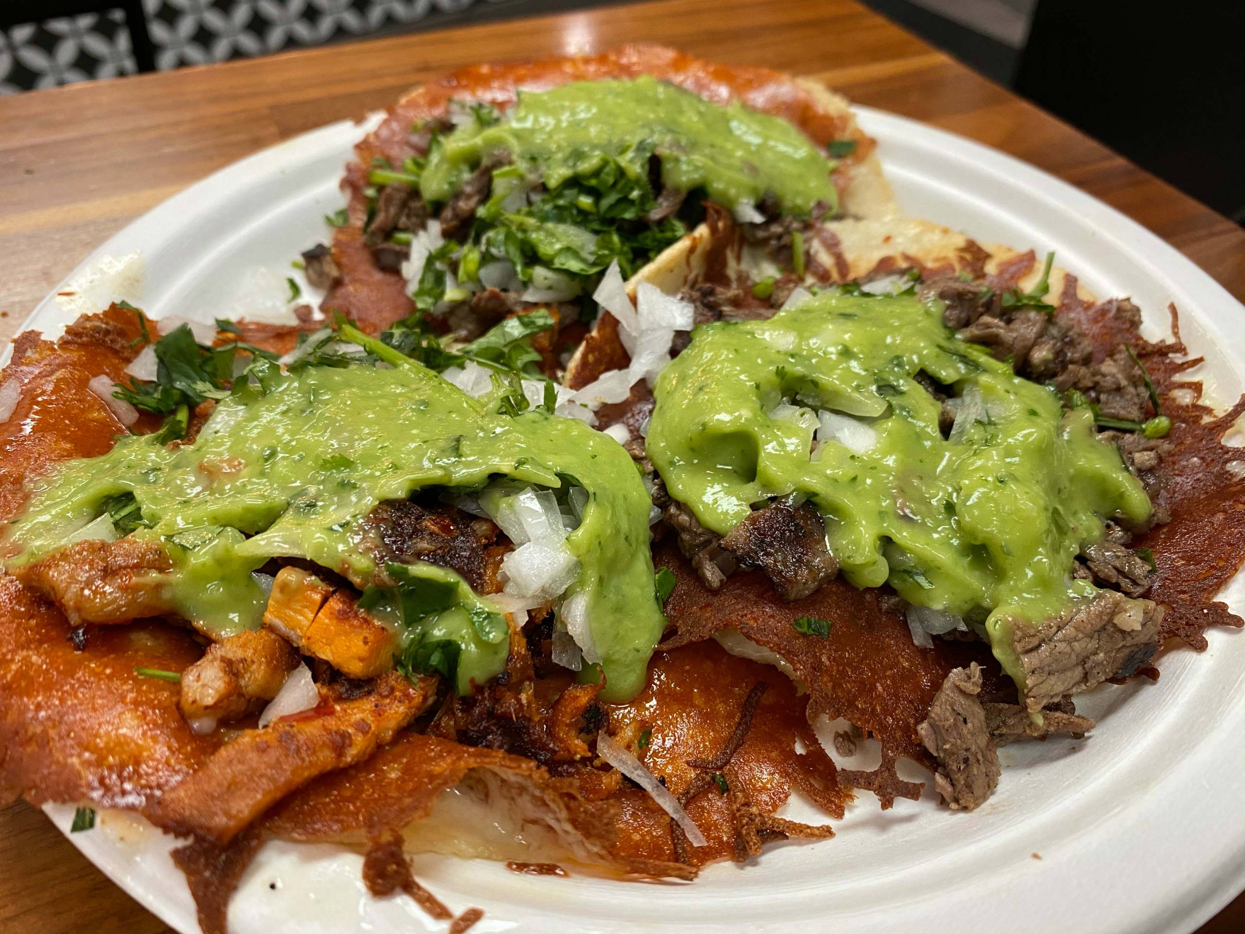 Three tacos, topped with guacamole and crispy cheese, on a paper plate.