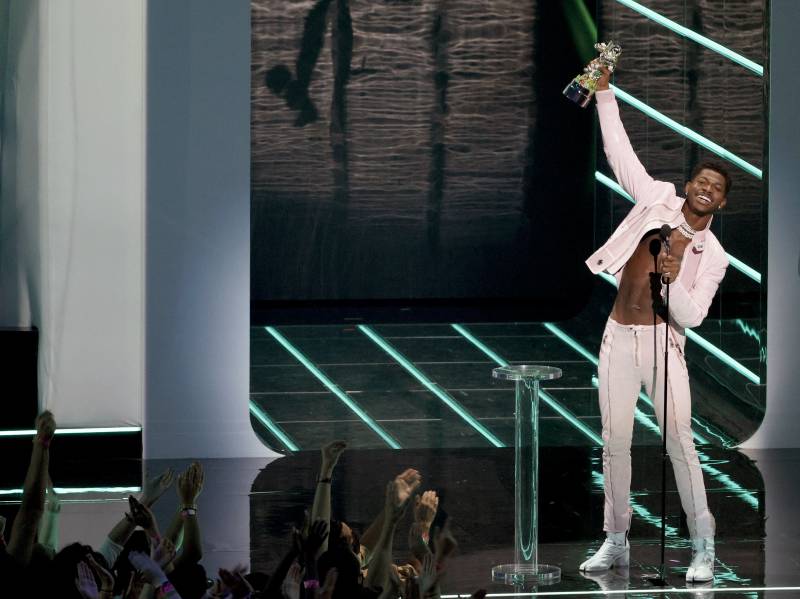 Little Nas X wearing white pants, white jacket and no shirt, smiles broadly and waves an award over his head.