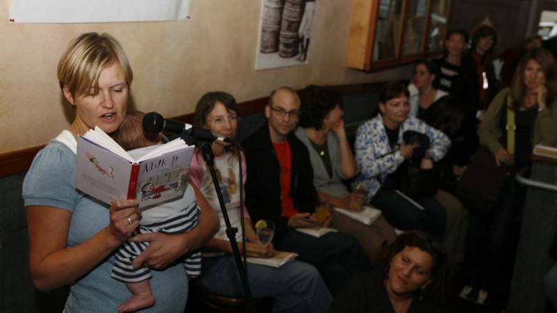A woman holding an infant reads from a book titled "Milk It" to an audience.