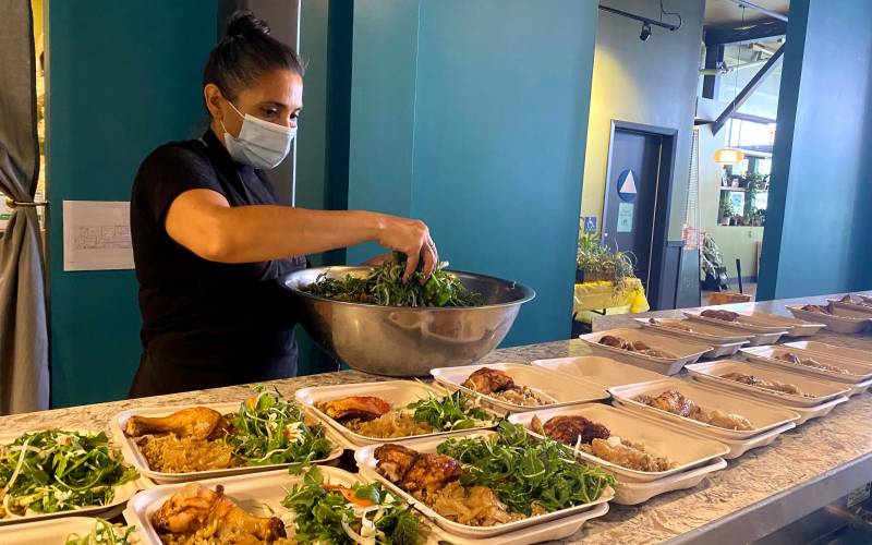 A woman wearing a face mask transfers portions of salad greens into takeout containers lined up on the counter.