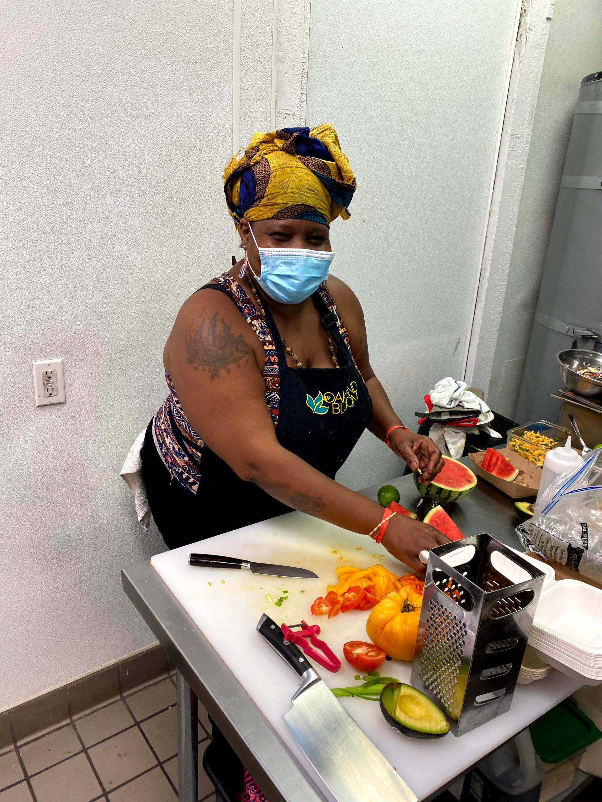 A Black woman wearing a face mask and black apron cuts up orange and red peppers on a small kitchen prep counter.
