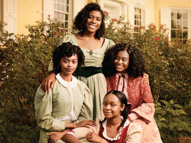 Four Black women pose together, smiling, in colorful 1860s period dresses, in a garden.