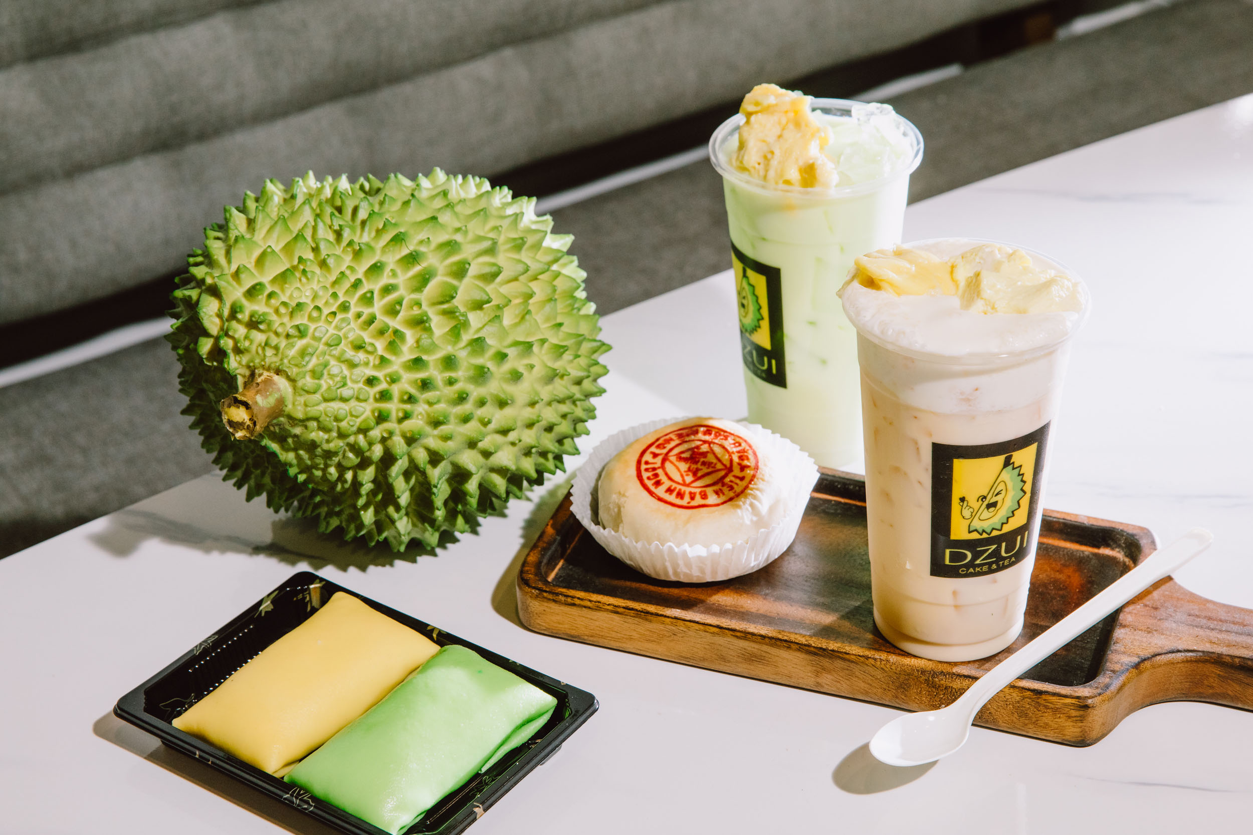 Two Vietnamese milk tea drinks, a pastry and a spiky green durian fruit spread out on a countertop.