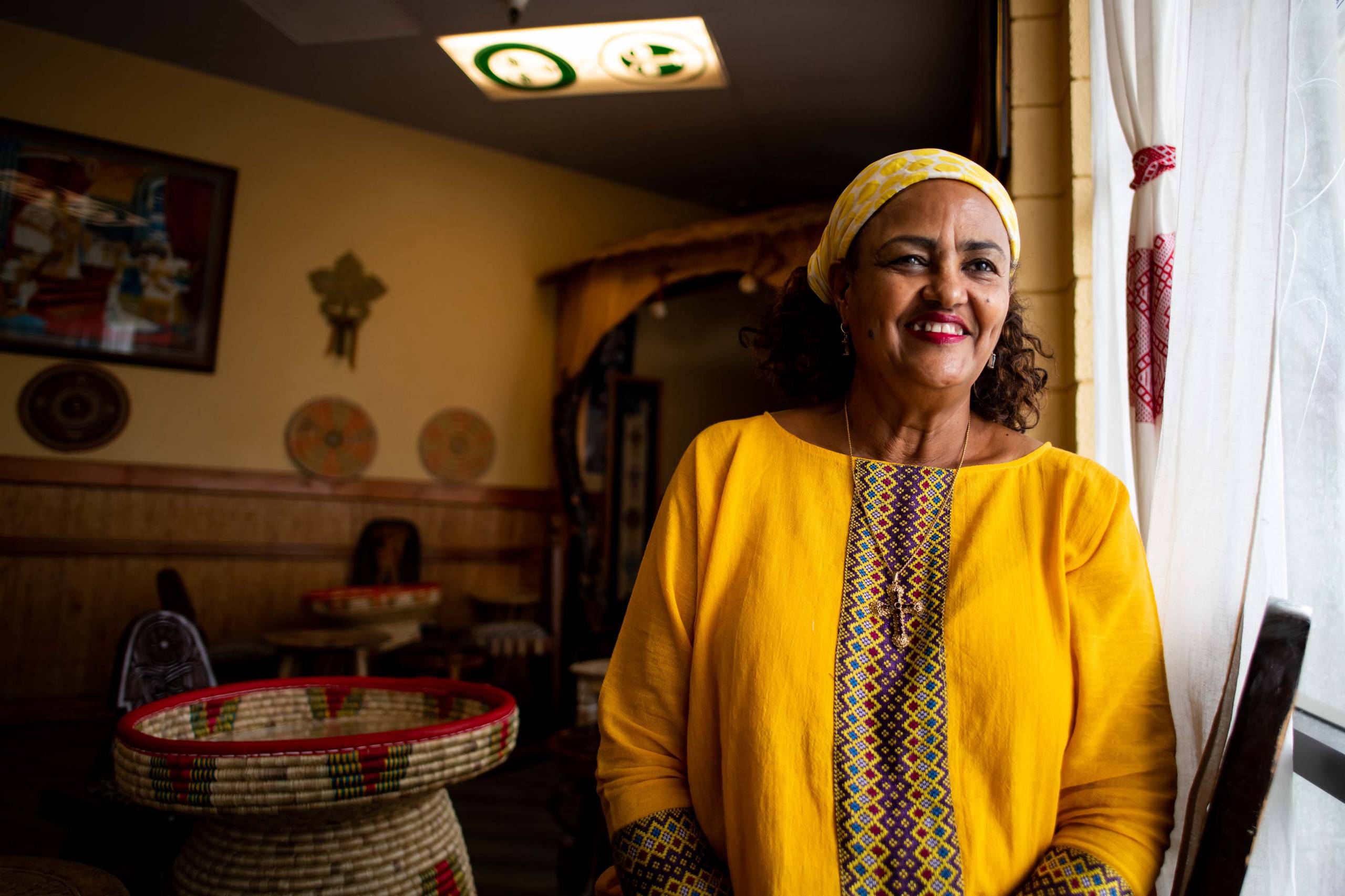 A woman in a bright yellow dress poses for a portrait inside her restaurant.