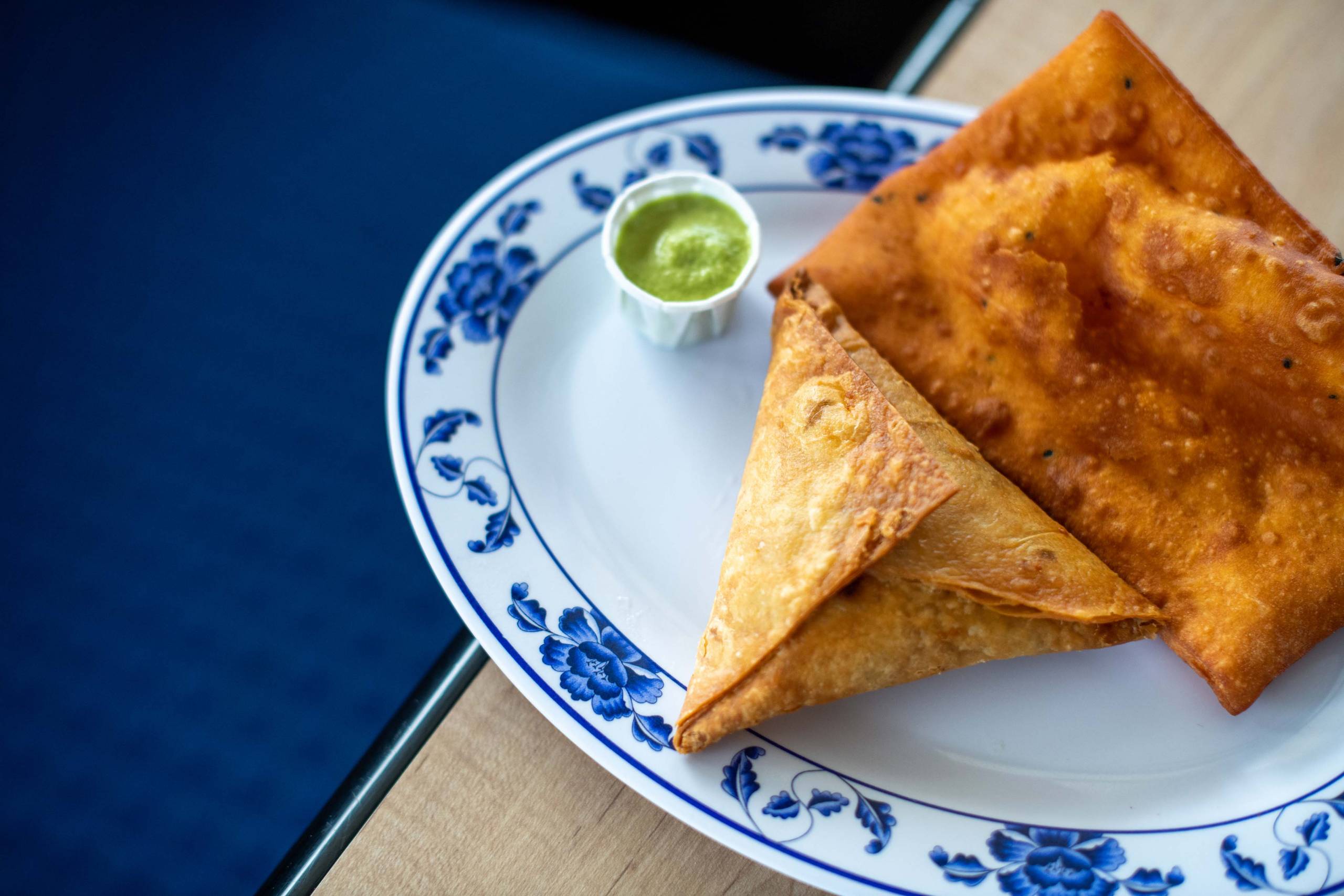 A samosa on a plate with a small container of green hot sauce.
