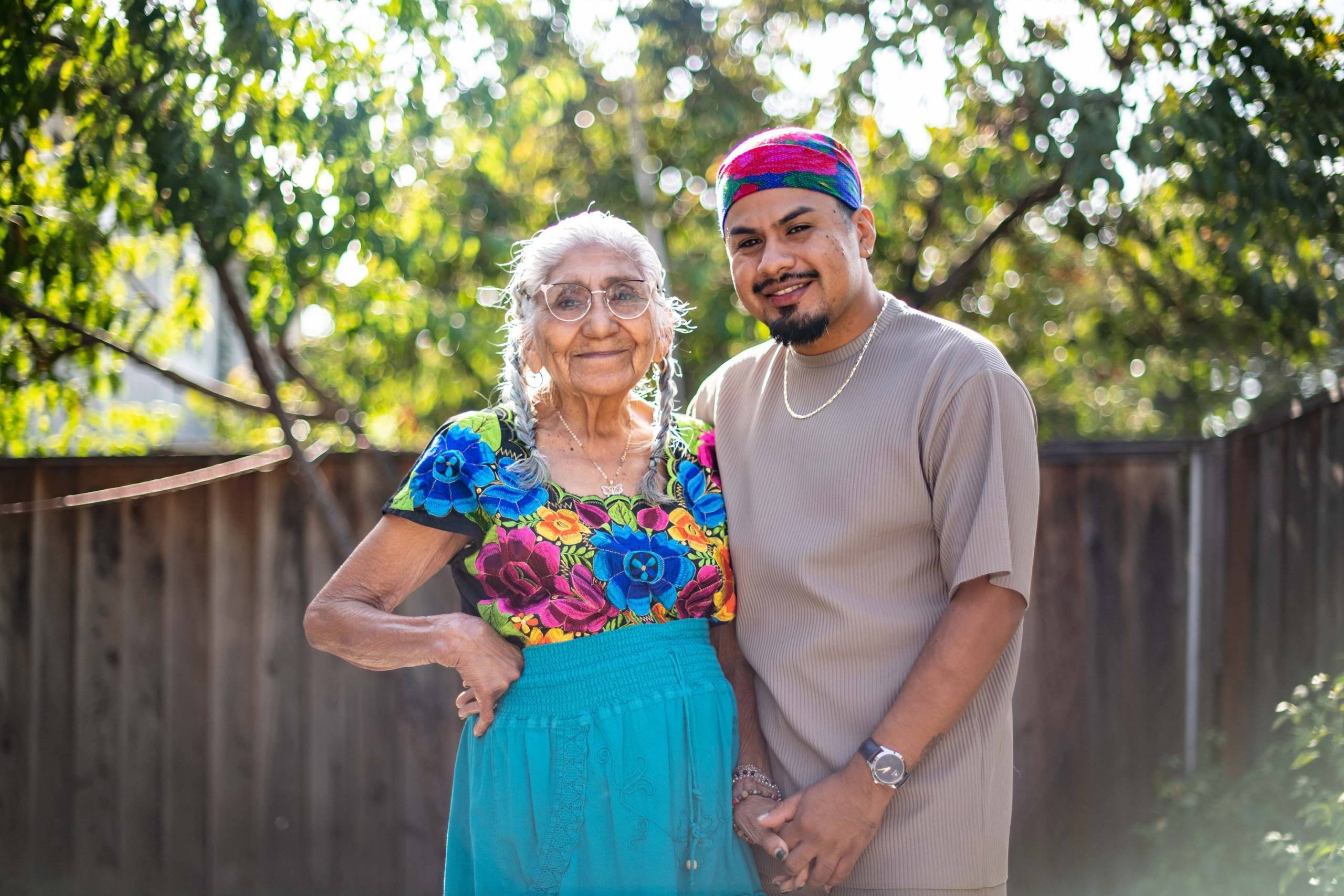 An elderly Latina woman and her grandson post for a portrait outdoors in their tree-lined backyard.