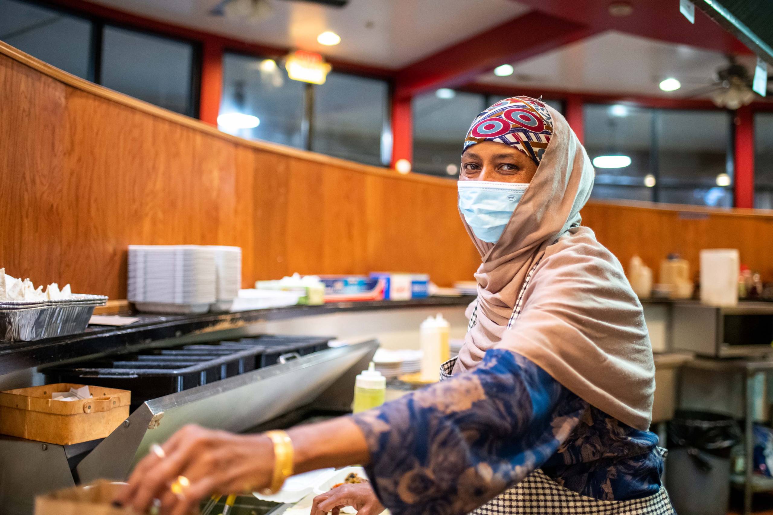 A woman wearing a face mask and a hajib prepares food over a flat-top griddle.