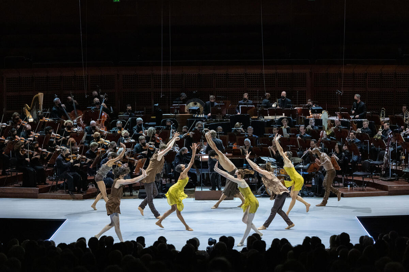 Male and female ballet dancers gather in a symmetrical formation in front of an orchestra.