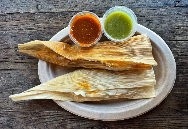 Two tamales in their corn husk wrappers on a paper plate, with red and green salsas on the side.
