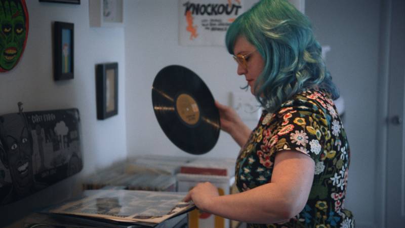 A blue-haired female DJ holds a record.