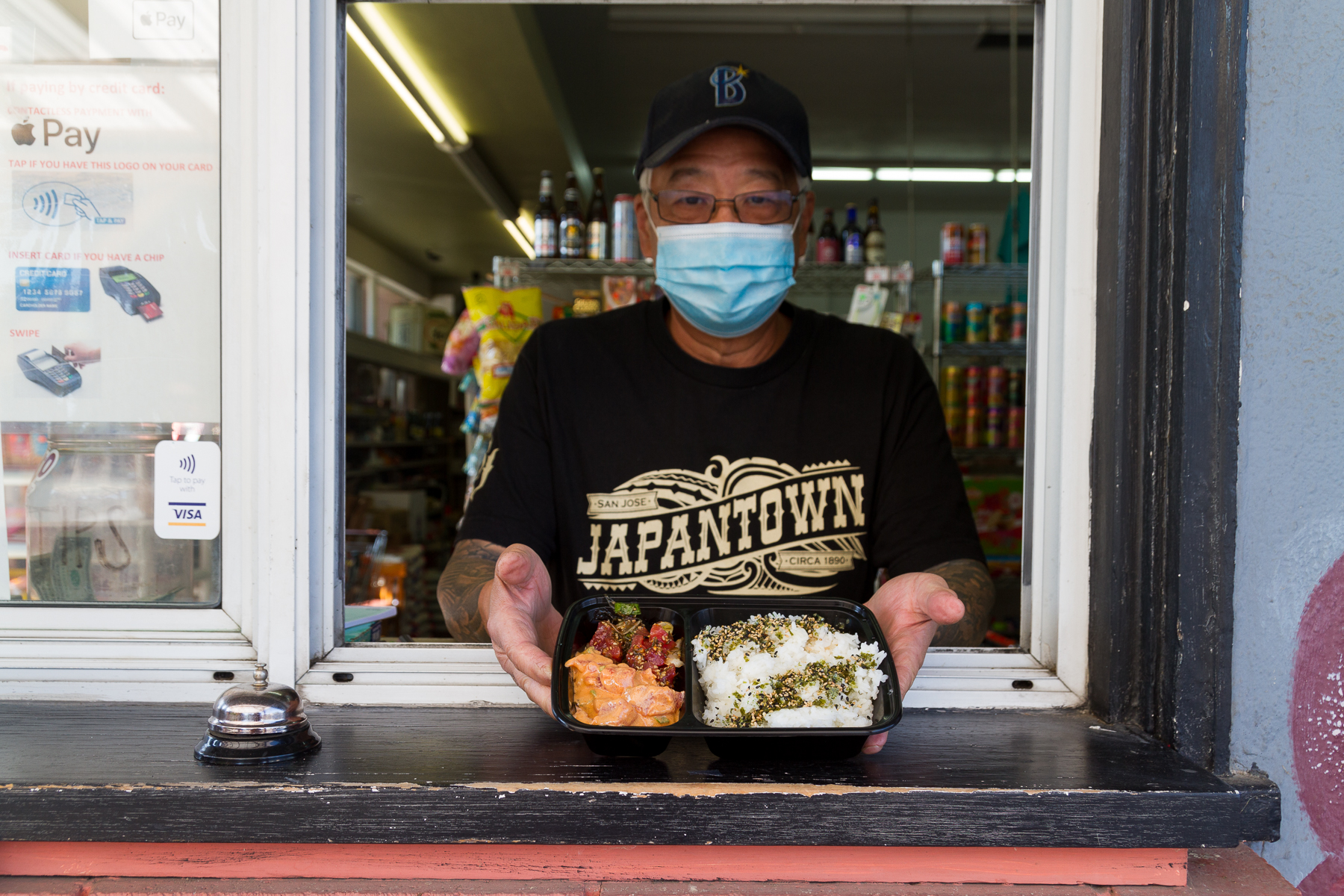 A man in a "Japantown" shirt holds out a tray of poke from the window of a Japanese market.