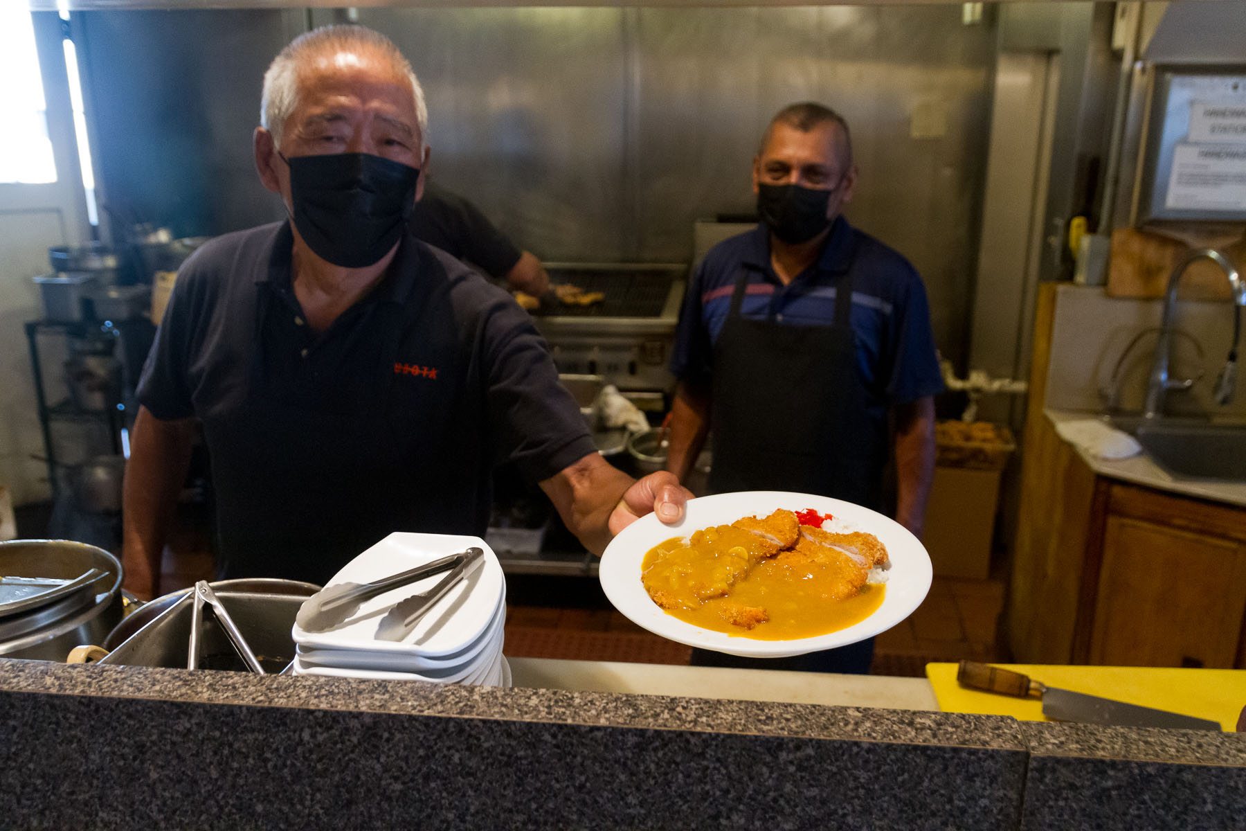 From inside a restaurant kitchen, a chef in a black uniform holds out a plate of katsu curry.